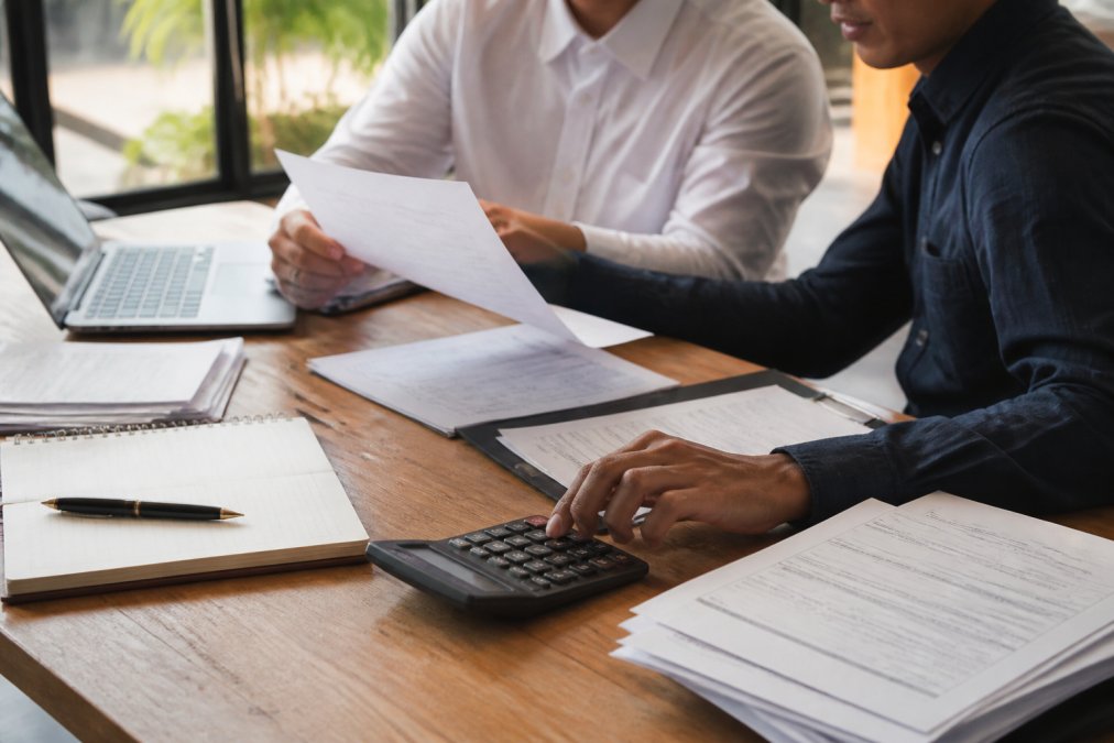A professional consultant and client reviewing printed manuscript pages together at a bright wooden desk, laptop open beside them, natural daylight from a nearby window
