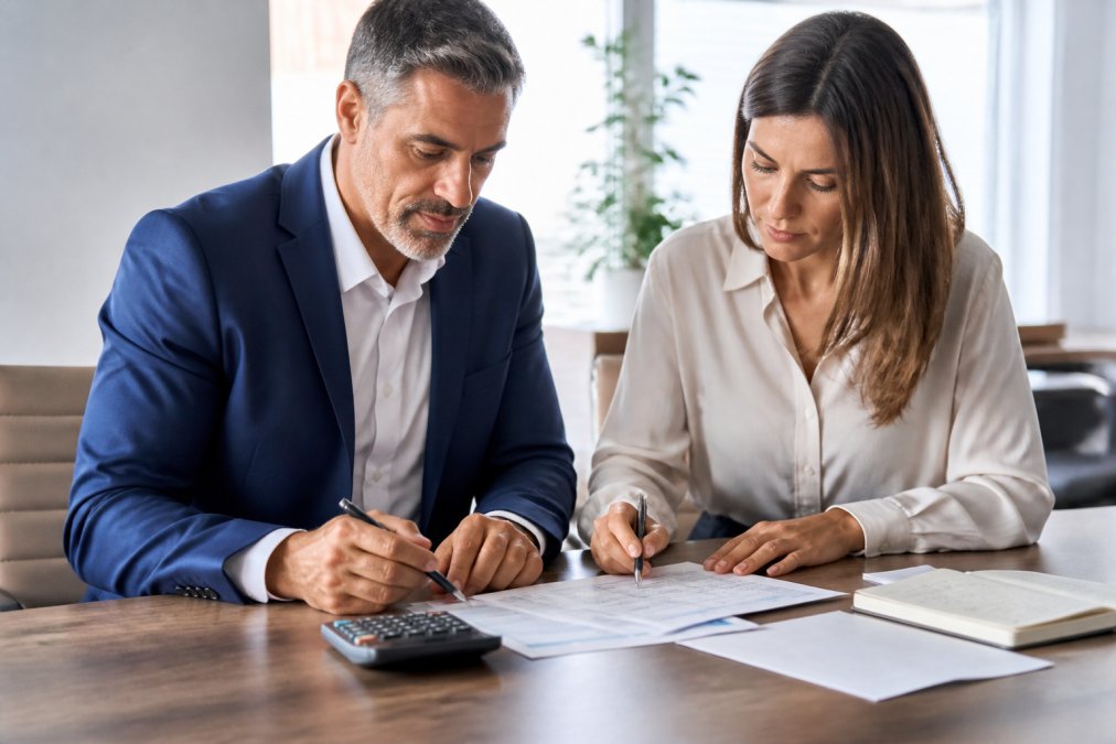 A business owner and professional advisor reviewing financial documents together at a polished desk, calculator and pen visible, bright professional office lighting