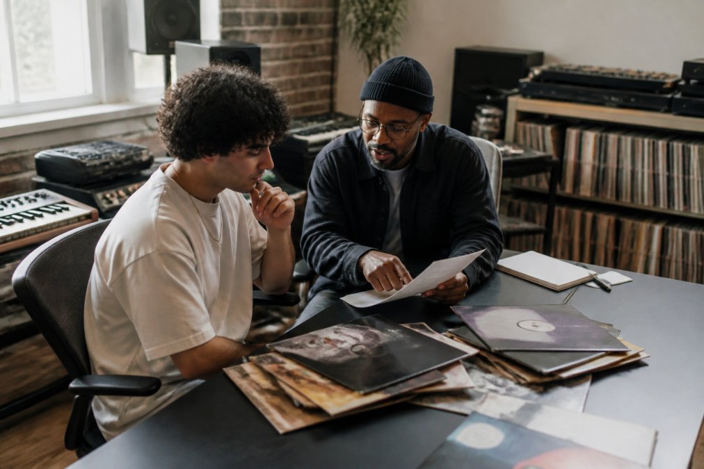 Professional A&R consultant reviewing artist materials and discussing creative strategy with a colleague in a modern recording studio office, with vinyl records and production equipment visible in soft natural lighting