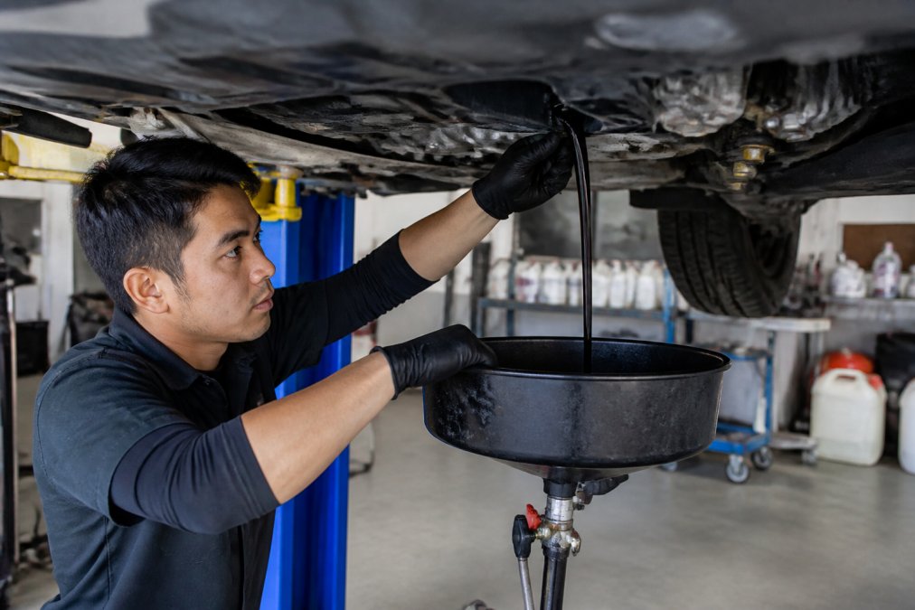 Professional technician performing engine oil and filter replacement on a Honda Civic, extracting dark used oil into a drain pan with service bay equipment and fluid containers visible in background