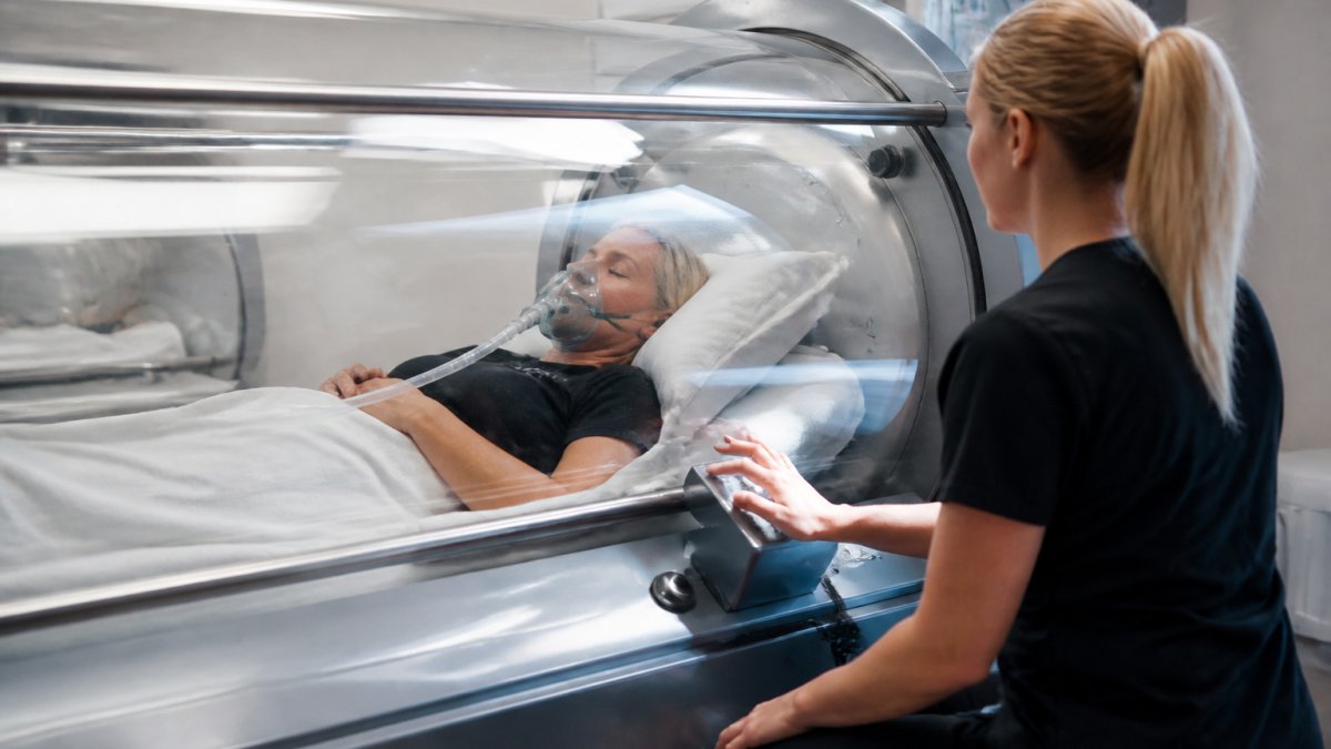Professional medical technician monitoring patient inside modern hyperbaric chamber with clinical monitoring equipment visible through transparent chamber wall