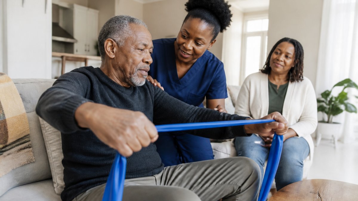 Licensed nurse assisting a senior with physical therapy during in-home skilled nursing in a bright living room.