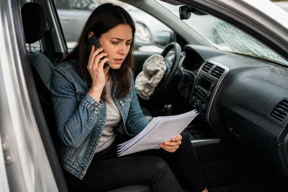 Person sitting in a damaged vehicle, on phone with concerned expression, reviewing accident documentation with paperwork visible on lap, natural daylight through car window