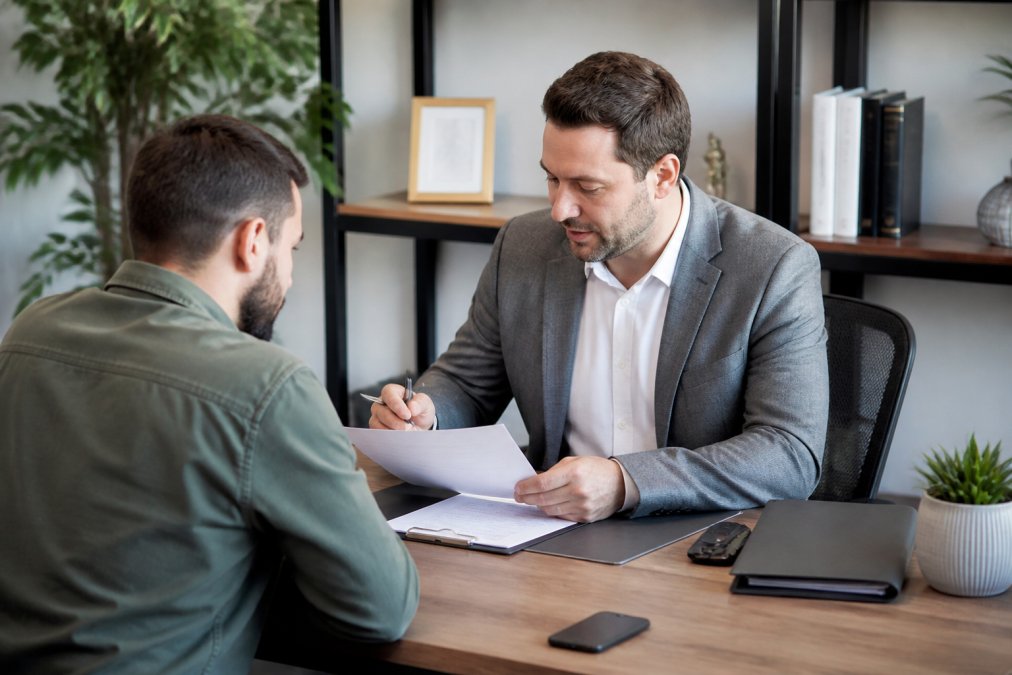 Professional advisor in modern office setting, seated at desk across from driver, reviewing accident claim documents together, calm lighting and organised workspace visible