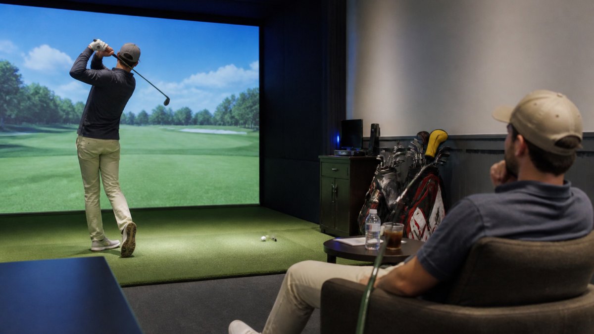 A relaxed beginner golfer mid-swing inside a modern indoor golf simulator bay, with a screen showing a fairway in front of them and a friend watching from a chair nearby