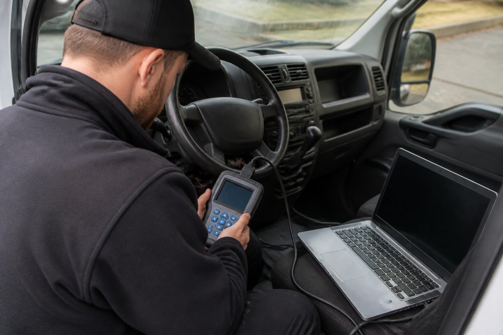 A locksmith in a mobile service van using a handheld key programming device connected to a vehicle's OBD port, with a laptop showing diagnostic software on the passenger seat