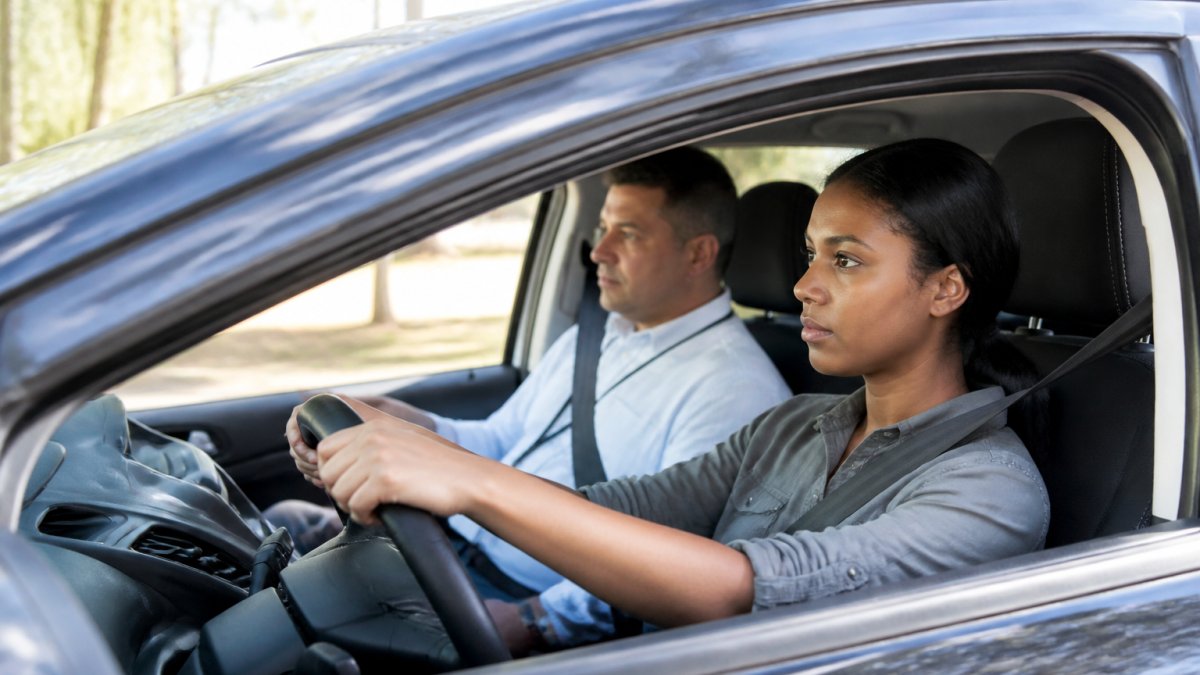 Professional driving instructor sitting calmly in passenger seat next to a focused adult learner gripping the steering wheel, both visible in profile through car window with clear road ahead and natural daylight