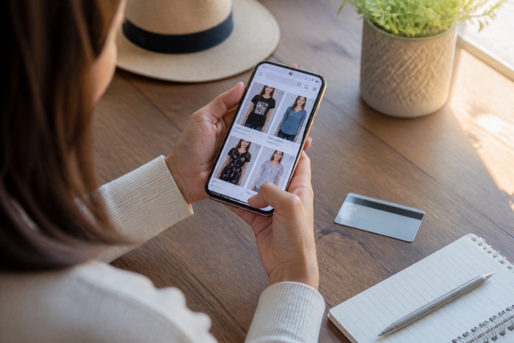 Close-up of a woman's hands holding a smartphone displaying an online clothing store product page, with a credit card and notepad nearby on a wooden desk under warm natural light