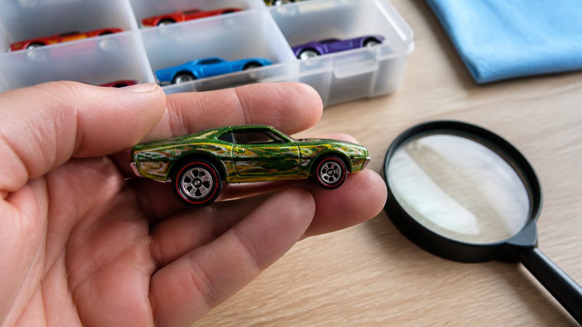 Close-up of a loose Hot Wheels Super Treasure Hunt car held in a collector's hand under bright light, showing Spectraflame paint finish and Real Riders wheels, with a magnifying glass nearby on a clean wooden desk