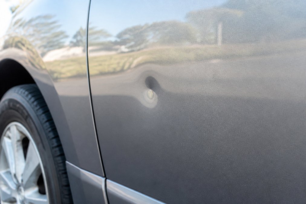 Close-up of a shallow minor dent on a car door panel, approximately one inch wide, showing intact paint finish and clean metal surface under natural daylight
