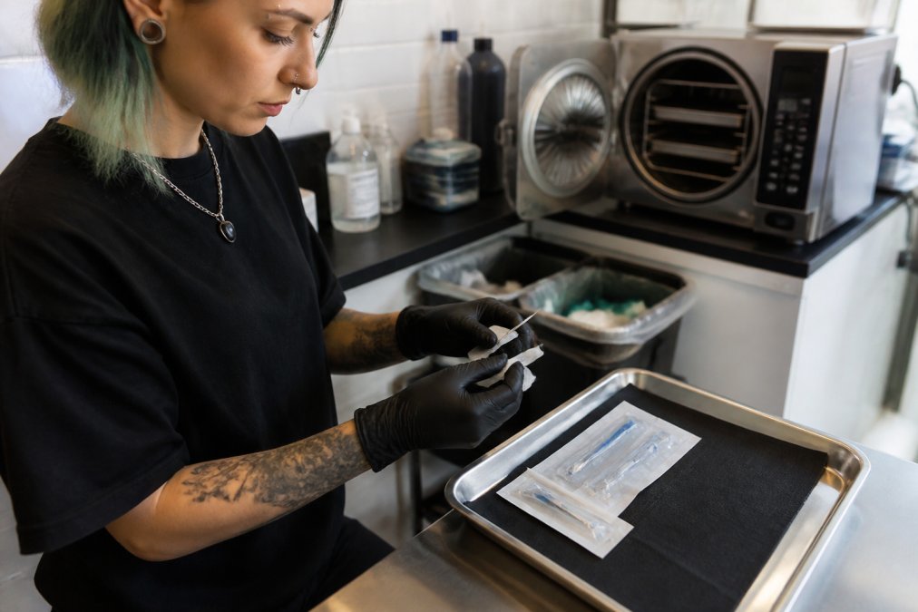 A professional piercer in gloves preparing a sterile, single-use needle on a clean tray in a bright, modern studio, with an autoclave visible in the background