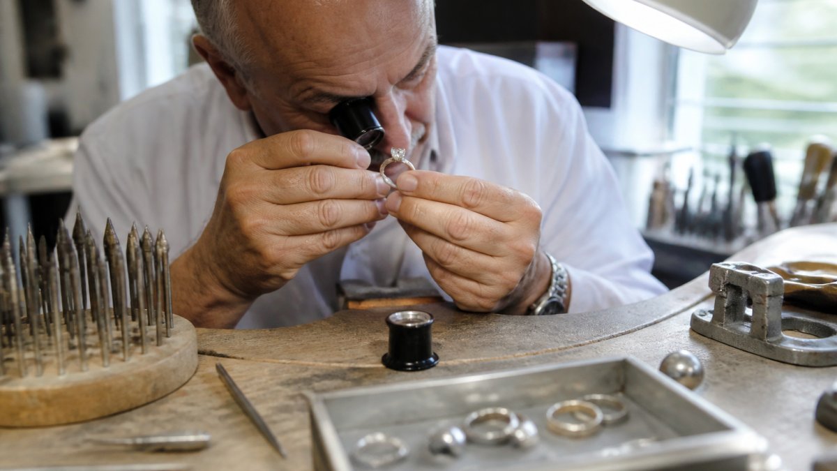 Professional jeweler using a jeweler's loupe to closely inspect a diamond ring's prongs and setting under bright overhead light, examining the metal-to-stone contact points