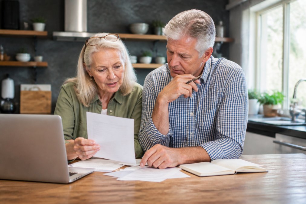 Mature professional couple reviewing retirement documents and financial projections at their kitchen table with a laptop and notepad, natural afternoon light streaming through windows