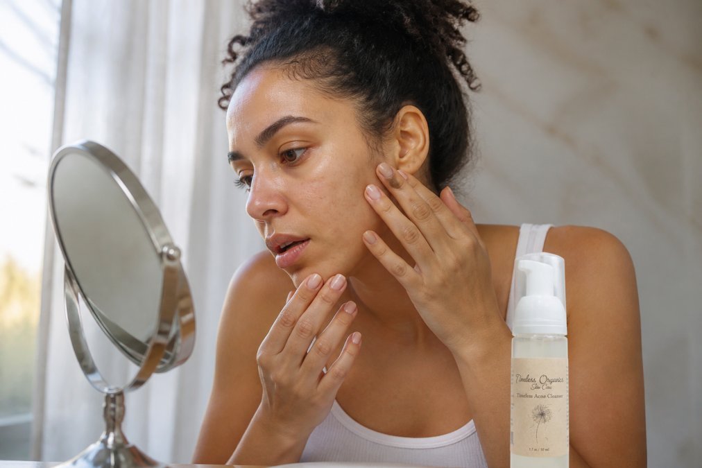 Person with diverse skin tone examining their face closely in natural daylight, showing different areas of skin texture and tone variation on cheeks and forehead