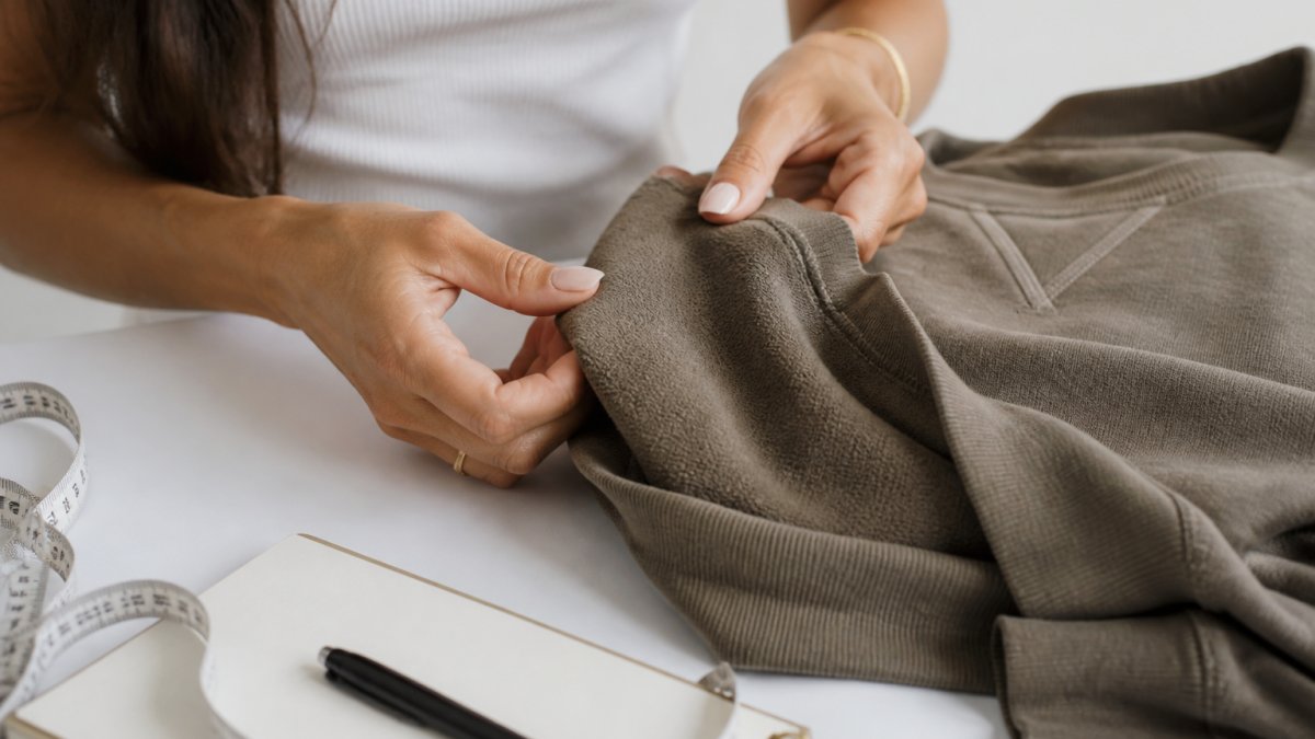 Close-up of a woman's hands examining the fabric and stitching of a soft knit sweatshirt on a clean white surface, with a measuring tape and notepad nearby
