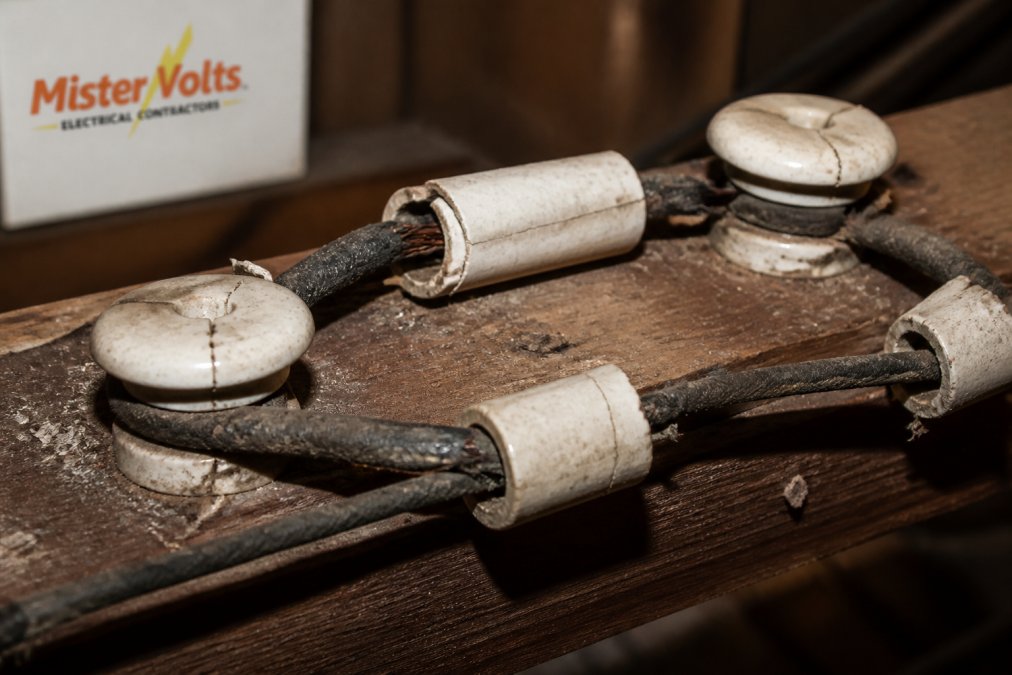 Close-up of deteriorating ceramic knob and porcelain tube insulation on aged copper conductors in an attic space, showing visible cracks, discoloration, and exposed wire sections