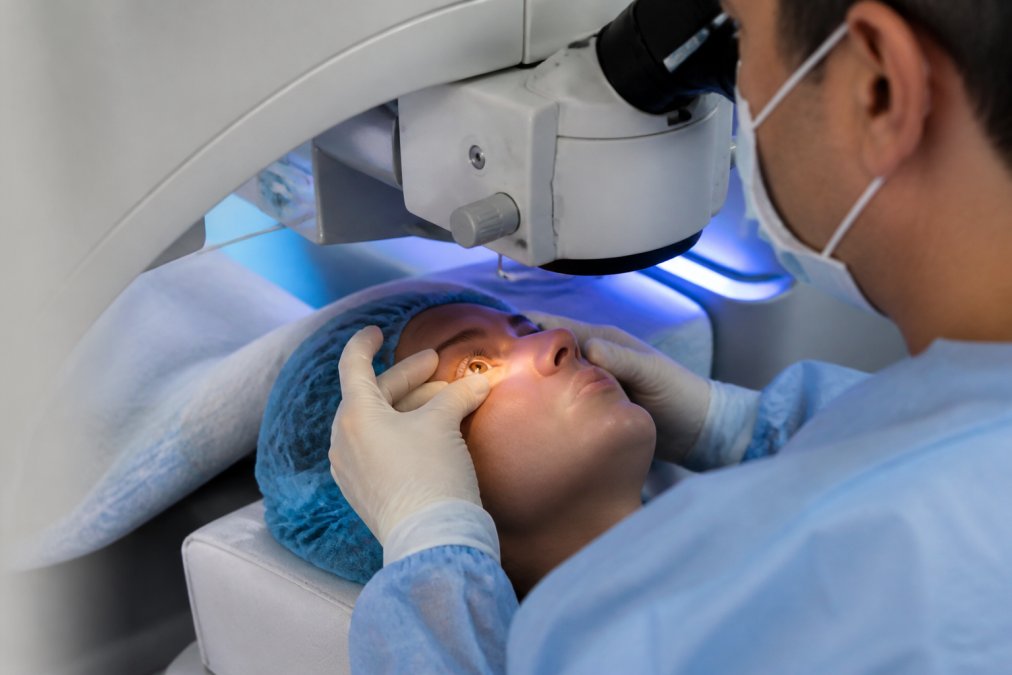 Ophthalmologist's hands positioning patient's eye under surgical laser apparatus in modern surgical suite with blue operating room lighting and precision equipment visible