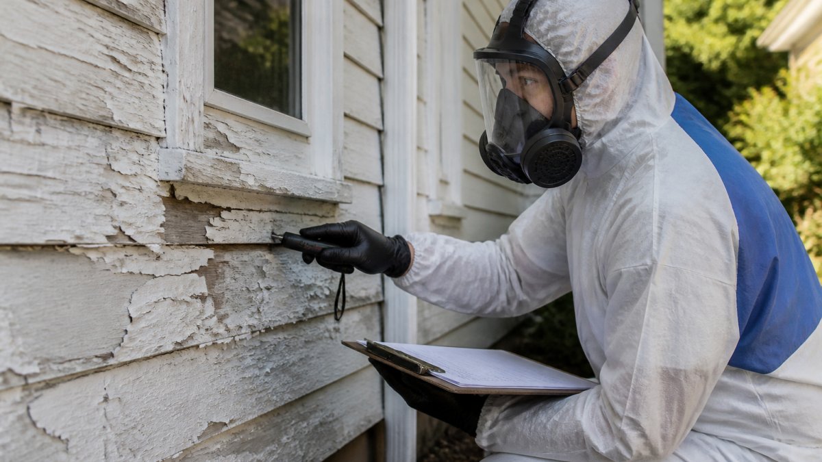 A certified inspector in protective gear examining peeling paint on the exterior of an older home, holding a clipboard and testing tool