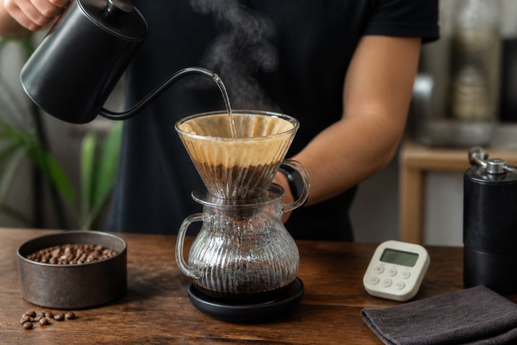 Pouring hot water over coffee grounds in a pour-over dripper with a kitchen timer and beans nearby