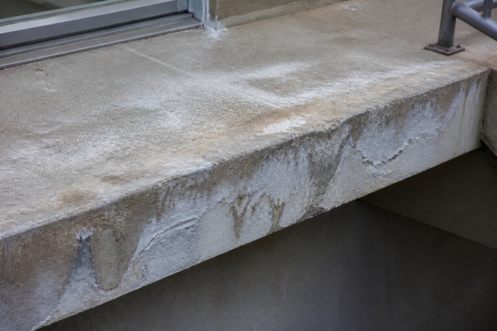 Close-up of concrete balcony surface showing visible water stains, efflorescence deposits, and early signs of deterioration with moisture damage visible on the edge