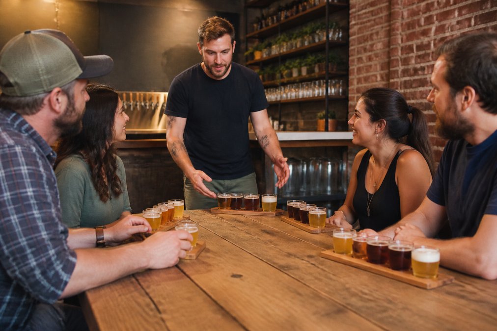 A group of adults seated around a wooden table in a warm, independent taproom, each with a beer flight of small glasses in front of them, while a host gestures toward the beers