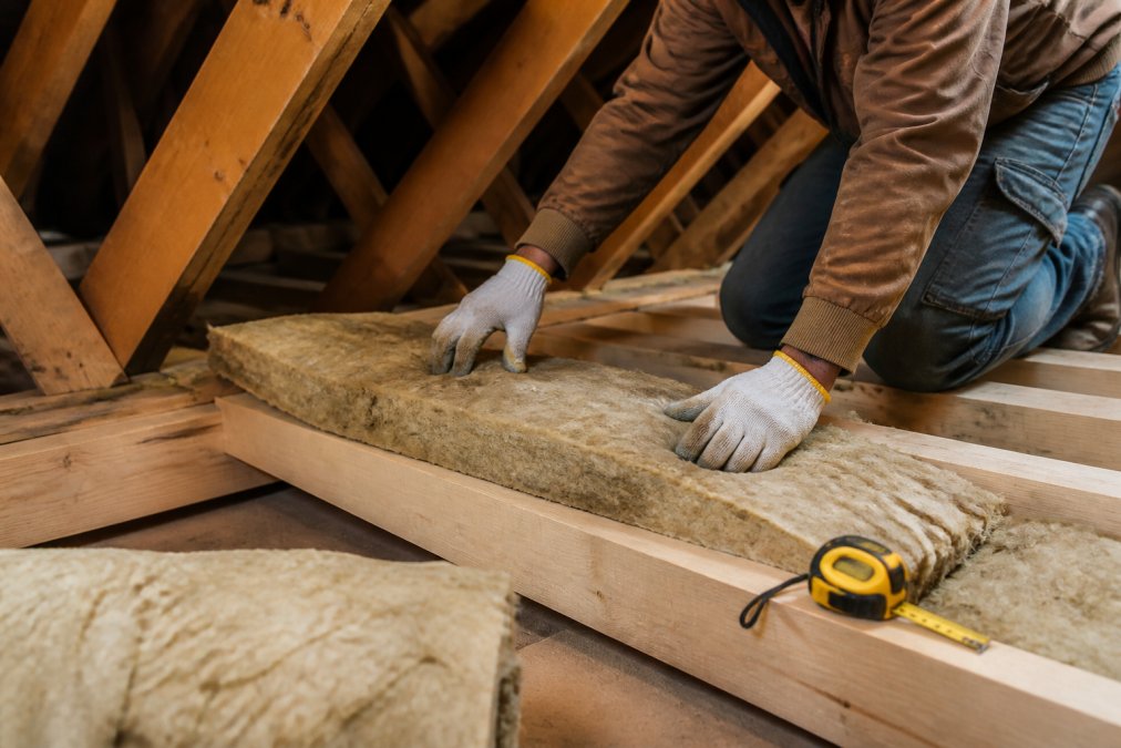 Close-up of a professional installer's hands laying mineral wool blanket insulation between wooden loft joists, with a yellow tape measure visible and dust motes in the daylight