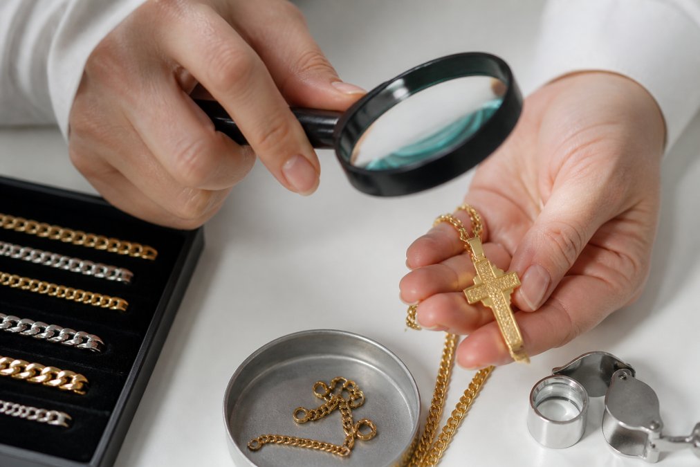 Close-up of hands examining a gold-plated cross necklace with a magnifying glass, showing clasp details and base metal composition against a clean white surface