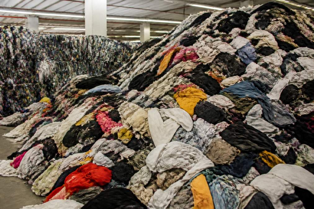 Piles of discarded textiles and unworn garments stacked at a sorting facility under fluorescent lighting, showing the scale of fast fashion surplus waste