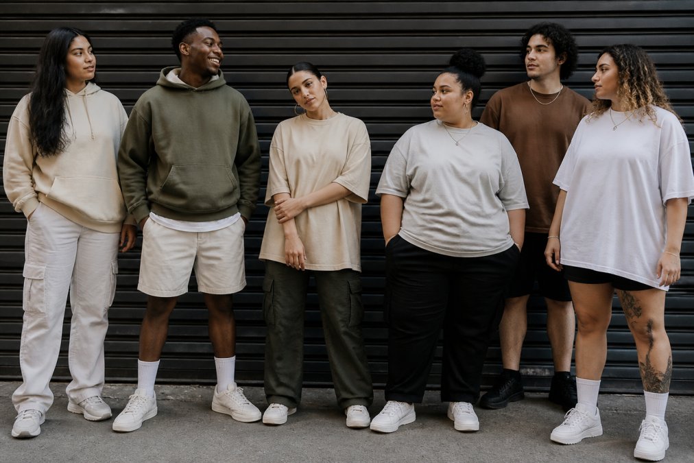 Diverse group of young adults wearing custom-printed streetwear in sizes ranging from 2XS to 6XL, posing confidently in urban setting with natural daylight, showcasing individual style and inclusive sizing