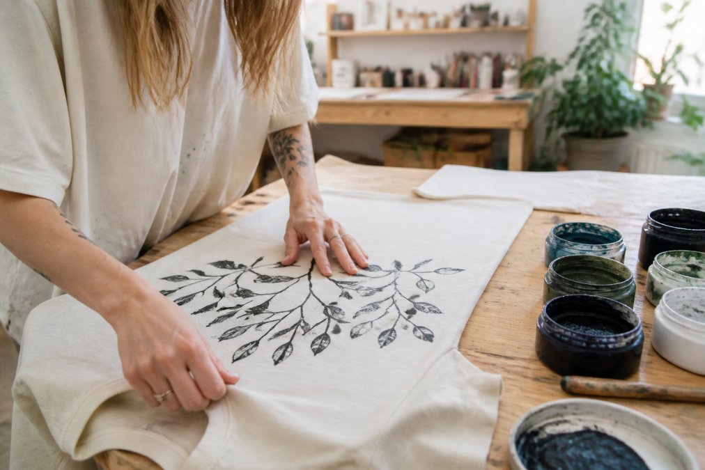 Hands carefully hand-printing a custom garment on organic cotton fabric using water-based inks on a wooden work table, showing the artisanal craftsmanship and attention to detail in a bright studio setting