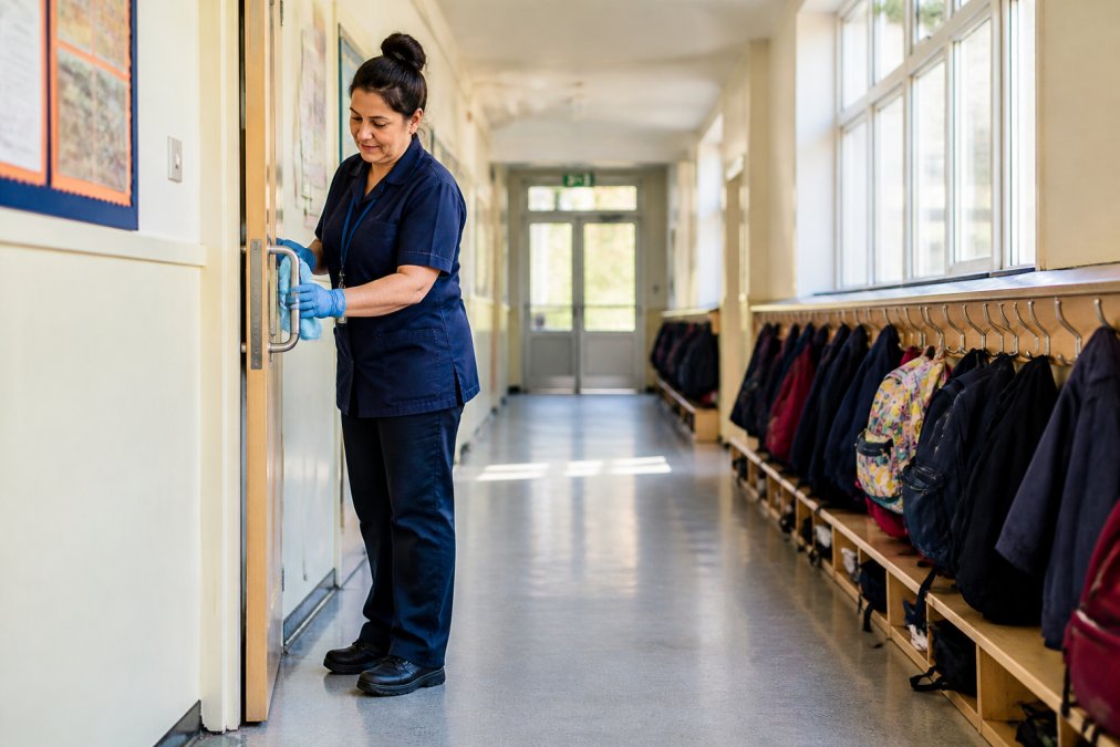 Professional cleaner methodically cleaning a primary school corridor with attention to detail around classroom entrances and high-touch surfaces, natural daylight from windows
