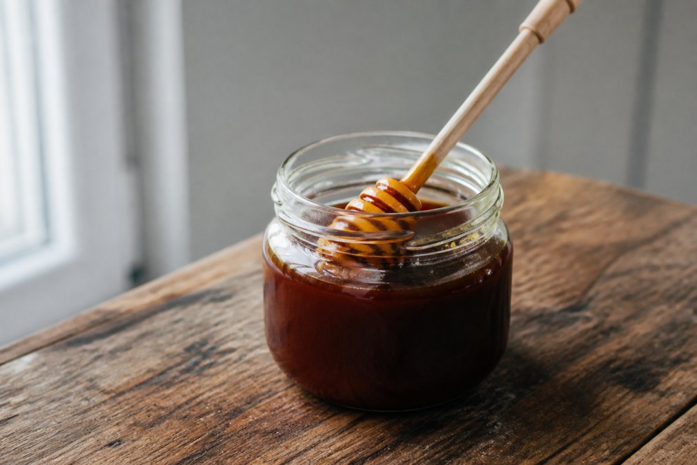 A clear glass jar of dark, thick Manuka honey on a rustic wooden table with a honey dipper resting inside, soft natural window light casting gentle shadows
