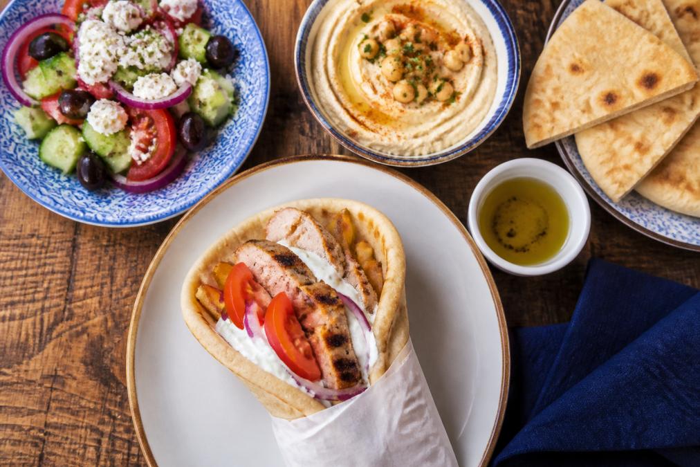 A colorful overhead spread of a Mediterranean-style table with a grilled chicken gyro, a Greek salad with feta and olives, hummus with pita, and a small bowl of olive oil for dipping in warm natural light