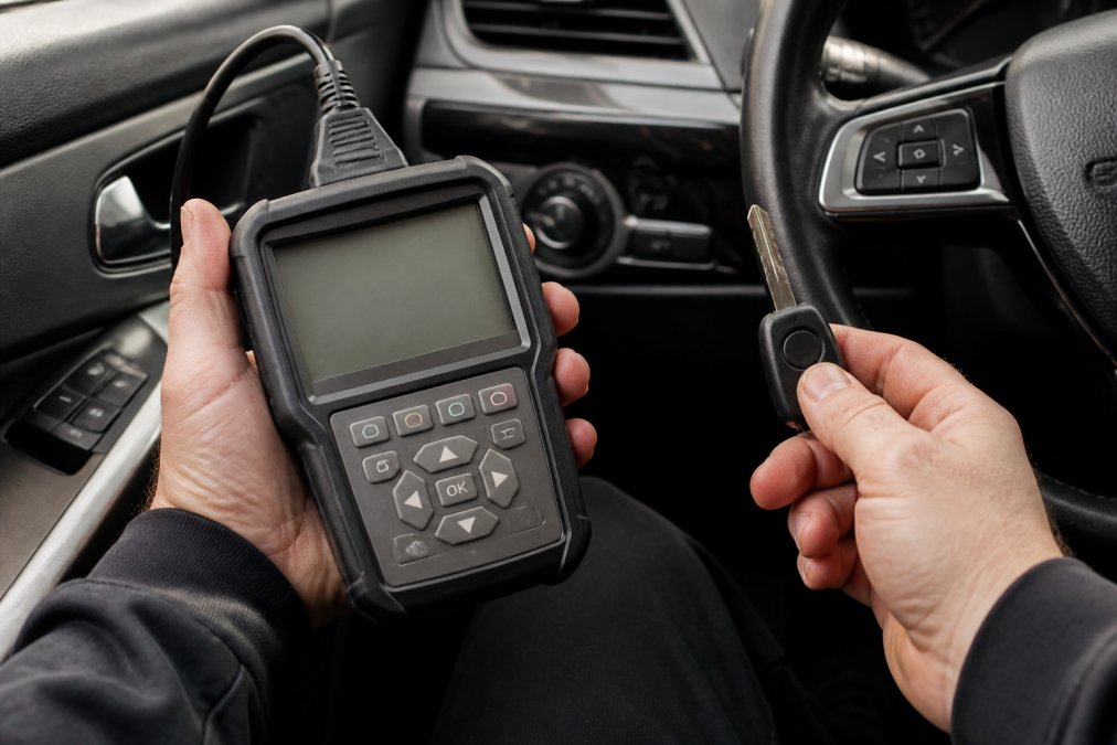 Close-up of professional locksmith's hands holding a transponder key and diagnostic scanner, showing precision tools and equipment required for safe key programming with vehicle interior visible in soft focus background
