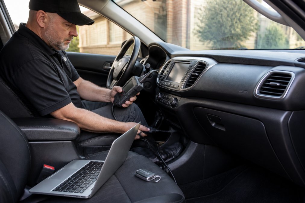 A locksmith technician seated in the driver's seat using a handheld programming device connected to a car's OBD port, key fob and laptop resting on the passenger seat, daylight through the windshield