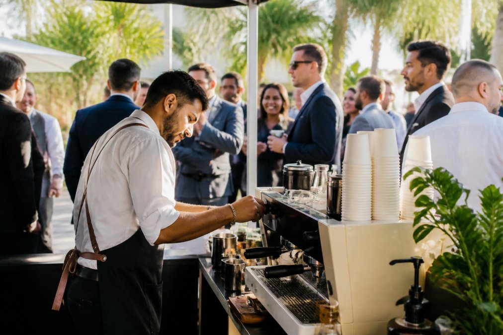 Professional barista preparing multiple espresso drinks at a mobile coffee cart during an event, with corporate guests visible in background holding branded cups