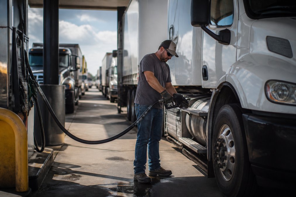 Commercial fleet truck driver refueling at a busy gas station pump during midday, showing the time and effort involved in traditional refueling with other vehicles waiting in background
