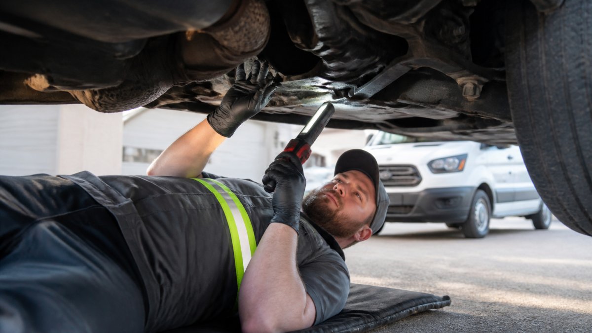 Professional mechanic in work gear inspecting the underbody and suspension of a vehicle using a portable light and diagnostic tools at a customer's driveway