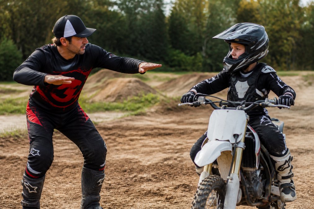 Young beginner motocross rider receiving instruction from a coach on a dirt track, with the coach demonstrating proper body positioning while the rider sits on a dirt bike wearing full protective gear and helmet