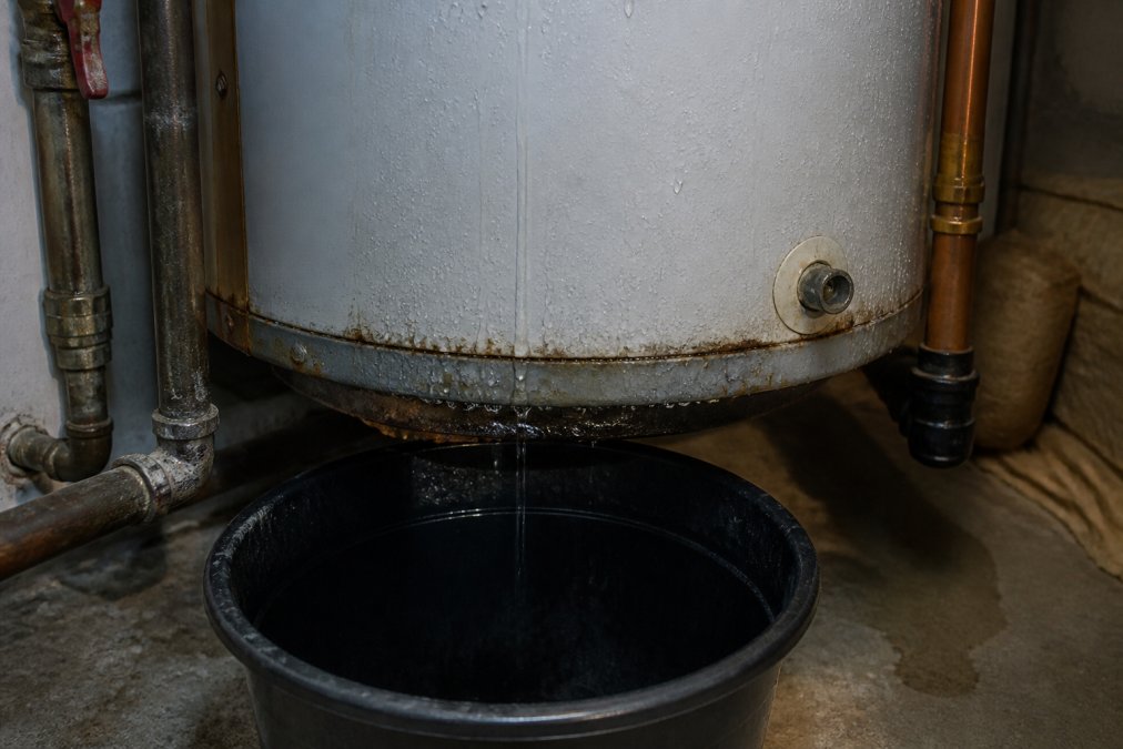 Close-up of water heater tank with visible water droplets and moisture on the lower tank section, a bucket positioned underneath to catch dripping water, basement lighting revealing the leak source