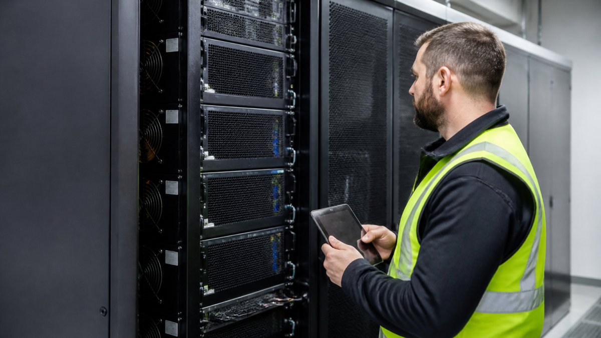 A data centre technician in a high-vis vest inspecting a modular UPS rack with multiple power modules, holding a tablet with monitoring software, cool blue lighting in a server room environment