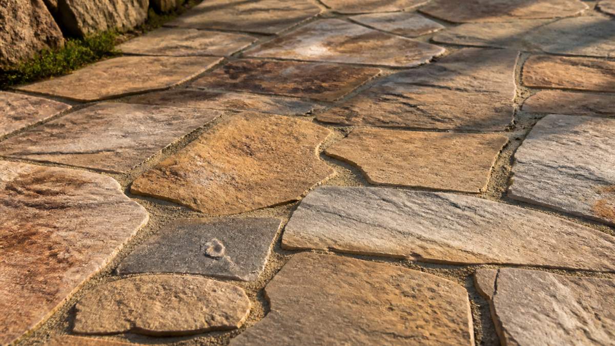 Close-up of a natural flagstone walkway showing varied earth-tone colors, irregular edges, and textured surface in warm afternoon sunlight casting subtle shadows across the stones
