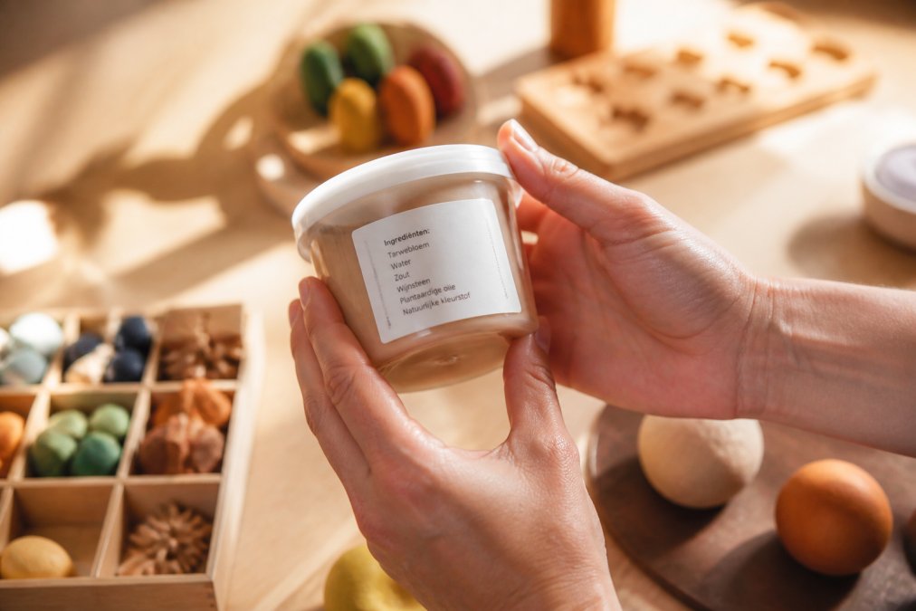 Close-up van schone handen van een volwassene die een potje natuurlijke speelklei vasthoudt met een duidelijk leesbare ingrediëntenlijst op het etiket, helder daglicht