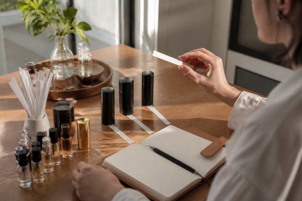 Fragrance enthusiast at home testing multiple niche fragrance samples on white wrist strips, with an open notebook and pen nearby for documenting scent impressions and longevity notes, natural window lighting streaming across the desk