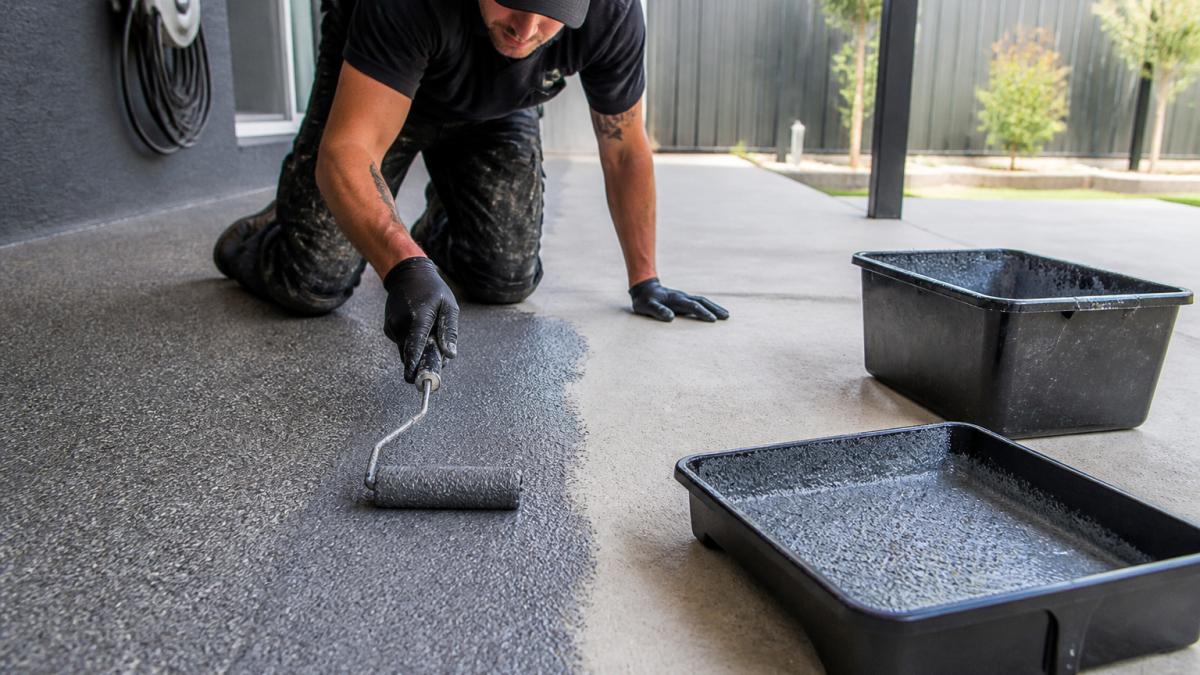 A close-up of a professional applicator rolling a textured non-slip coating onto an outdoor concrete patio, gritty finish visible, safety gear worn, natural daylight