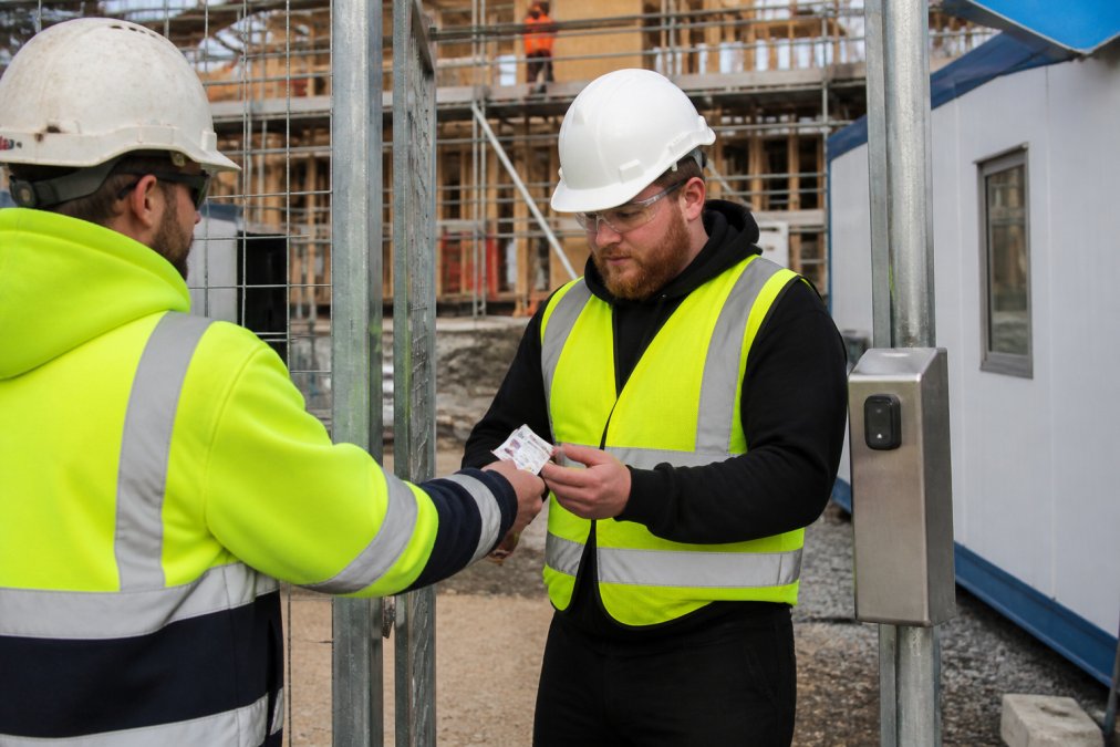 A construction site supervisor in a high-vis vest and hard hat checking a worker's white card on a tablet at a site entry gate, with scaffolding and a site office visible in the background