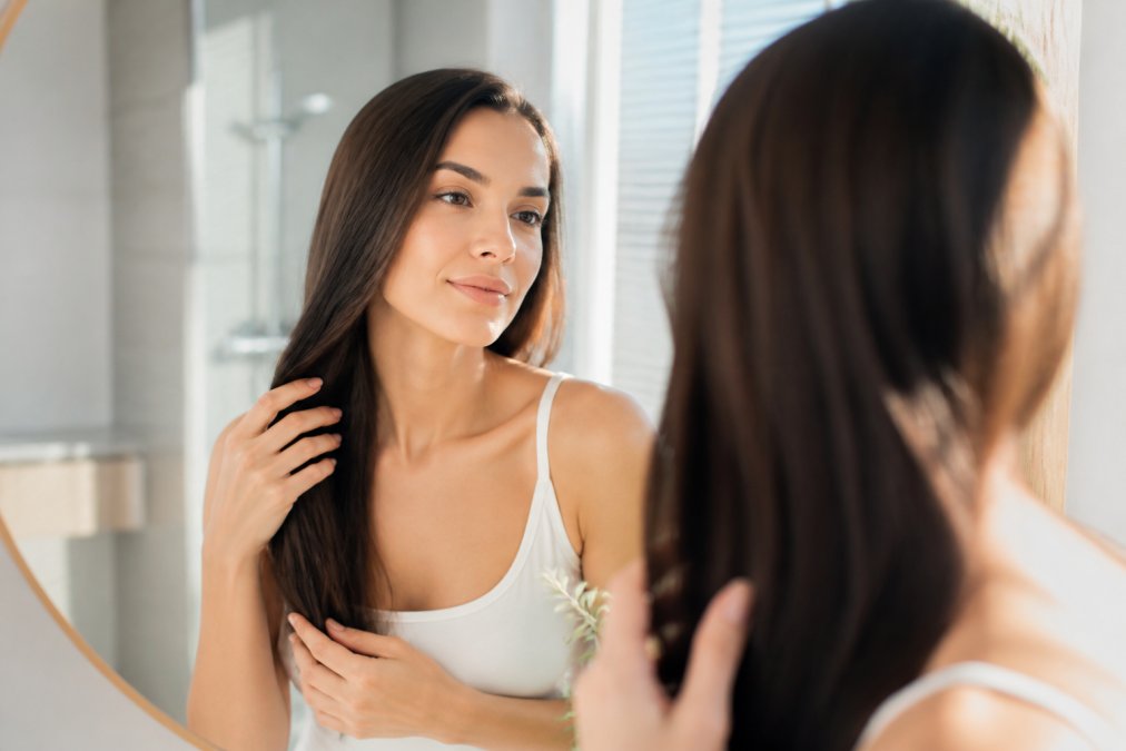 Close-up of a woman with healthy, shiny hair touching her smooth strands in soft natural light, her reflection visible in a mirror