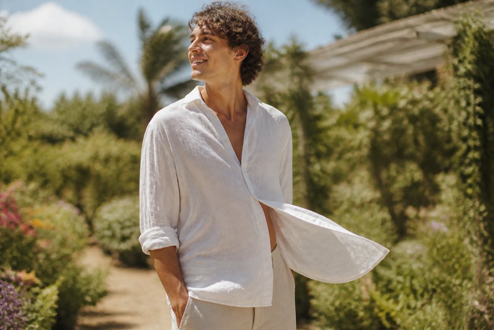 A person wearing a light-colored, flowing organic linen shirt standing in bright sunlight, with the fabric gently lifting in a breeze against a blurred summer garden background