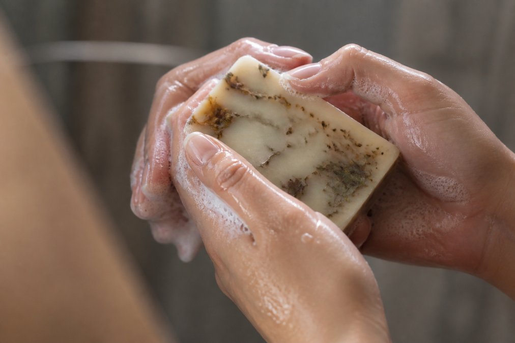 Close-up of hands holding a bar of organic soap with visible natural botanical ingredients, water droplets on skin showing moisture retention, soft lighting emphasizing creamy lather texture