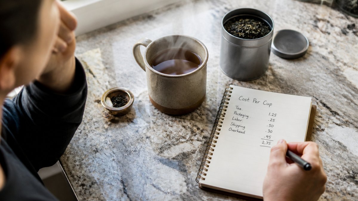 Overhead shot of a kitchen counter with a steaming mug of tea, a small tin of loose leaf tea, and a notebook with a cost-per-cup tally written by hand